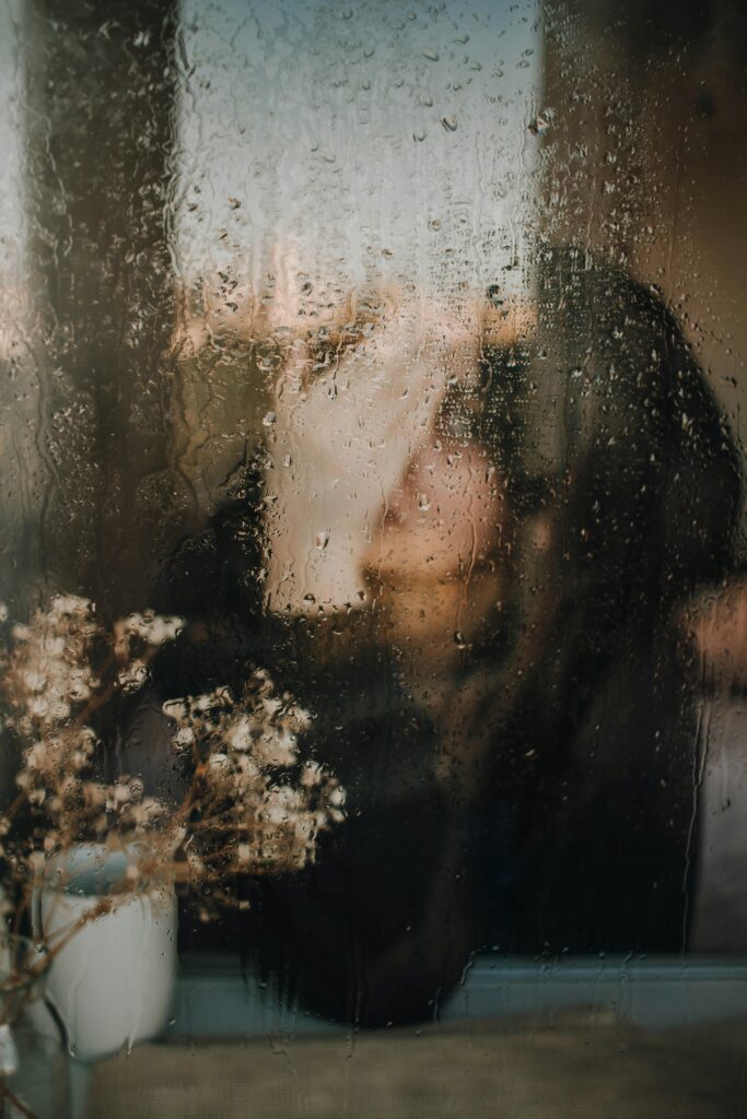 A woman sits behind a rain-streaked window holding a cup, creating a moody, introspective scene.