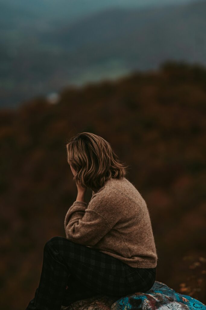 A woman sits on a hilltop in deep thought, surrounded by serene mountain views.