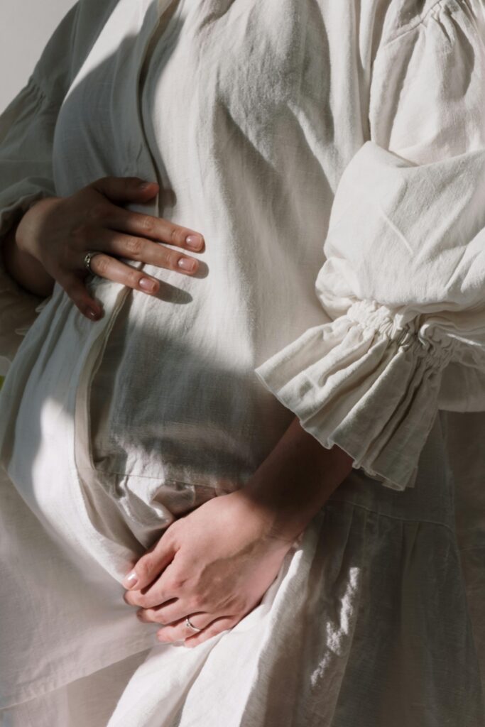 A pregnant woman gently cradles her baby bump in a sunlit white dress, showcasing elegance and maternal grace.
