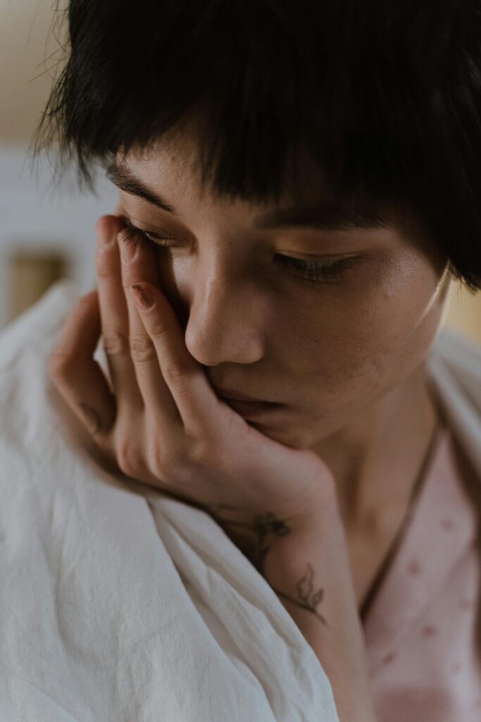 A young woman with short hair deep in thought, wrapped in a blanket indoors.