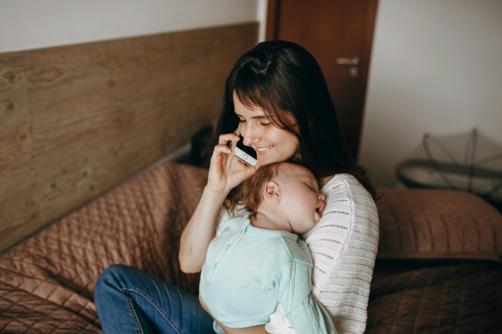 A smiling mother holding her sleeping baby while talking on the phone in a cozy bedroom setting.