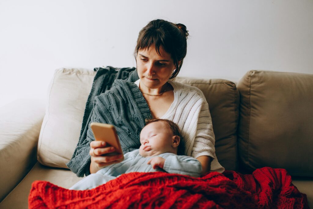 A serene moment with a mother browsing her phone as her baby sleeps on the couch.