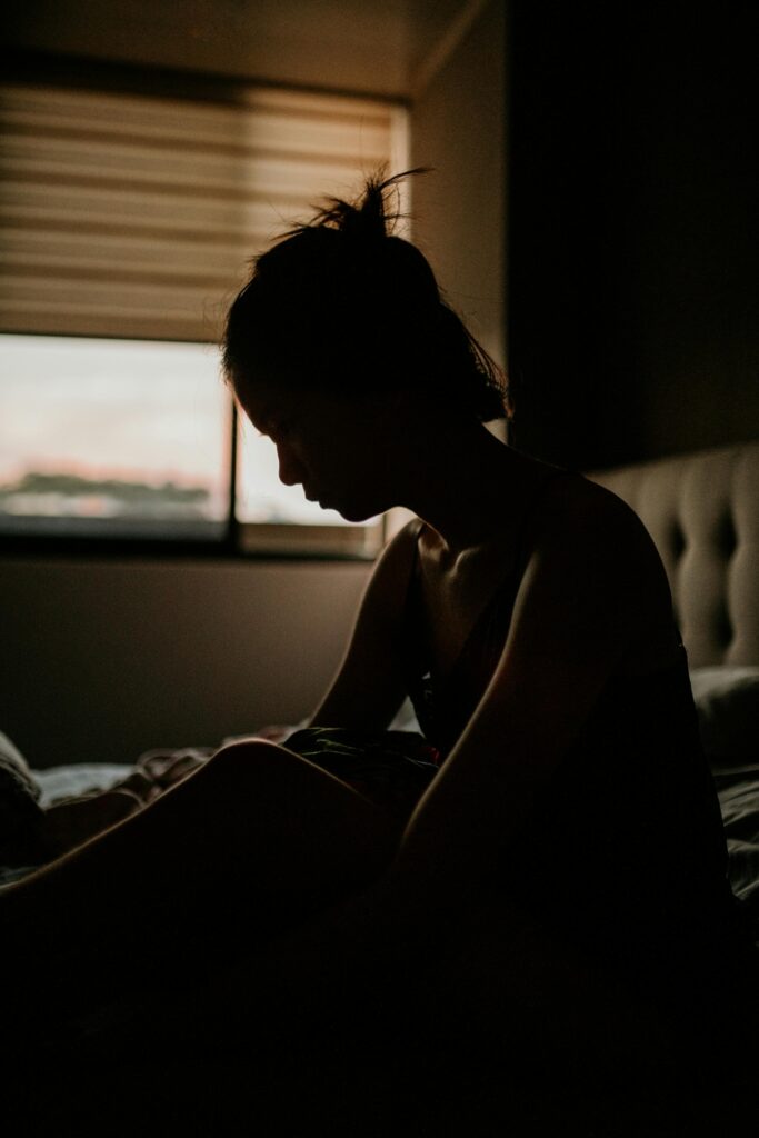 Silhouette of a woman sitting on a bed in a dimly lit room during early morning reflections.