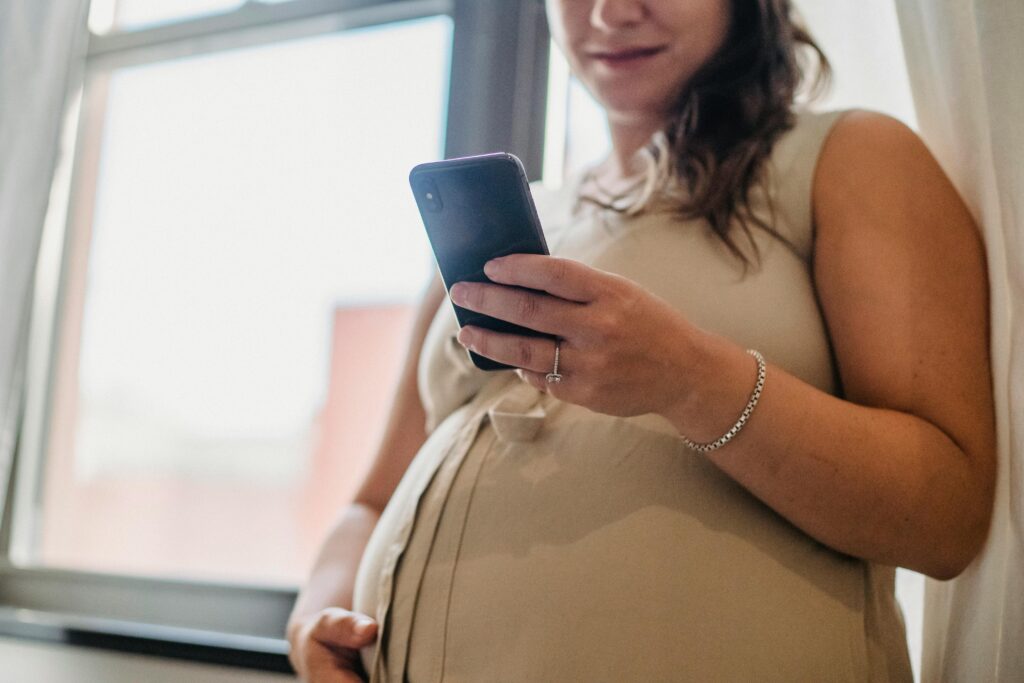 From below of unrecognizable pregnant female standing near window in room with cellphone and checking notifications in daytime