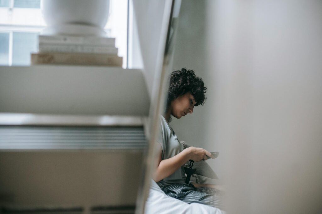 Side view of crop female text messaging on cellphone while resting near pile of books in house