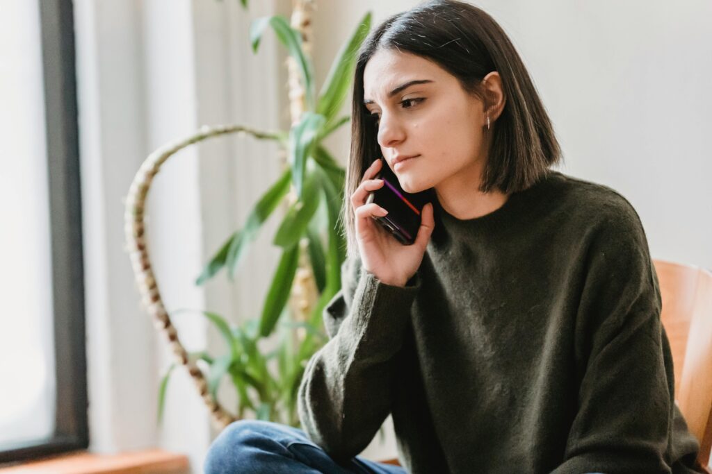 Calm young ethnic female millennial with dark hair in casual clothes having phone conversation while sitting on comfortable armchair at home in daylight